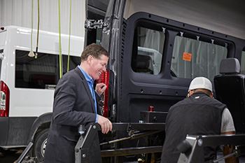 A smiling man in a suit is watching as a worker fixes the back of a van